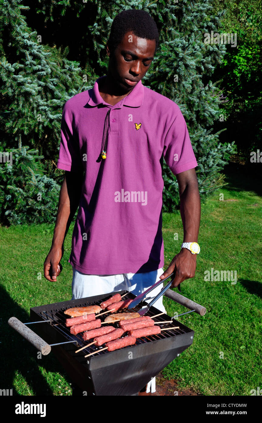Un bel, nero ragazzo adolescente servendosi di un paio di pinze per girare la carne in cottura su un barbecue a carbone in un giardino. Foto Stock