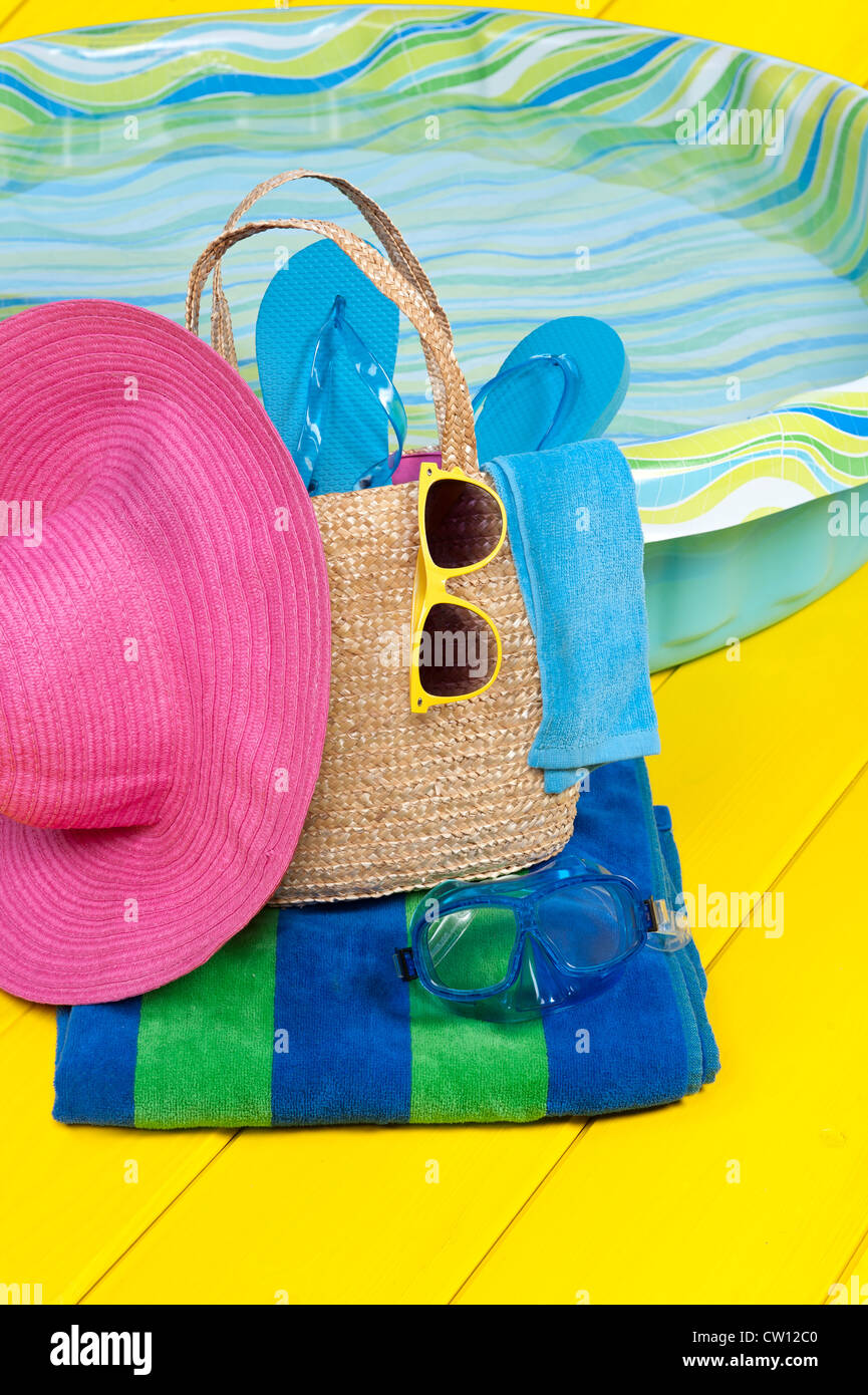 Una paglia borsa da spiaggia piena di accessori poggia su un telo da spiaggia accanto a un parco giochi per bambini Piscina in plastica piscina. Foto Stock
