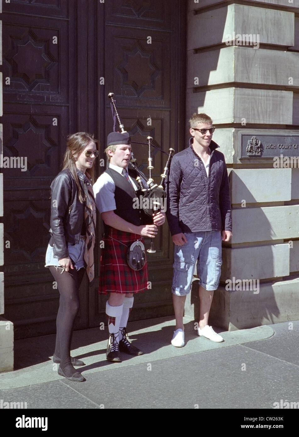 Giovani caucasici coppia turistica con Bagpiper musicista di strada al di fuori della Alta Corte di Justiciary, LAWNMARKET, Edimburgo, Scozia, Regno Unito Foto Stock