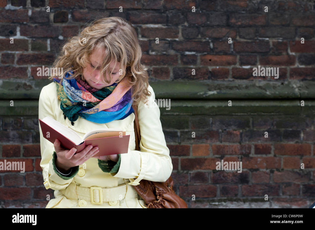 Giovane donna in un cappotto seduto davanti a un rosso un muro di mattoni la lettura di un libro Foto Stock