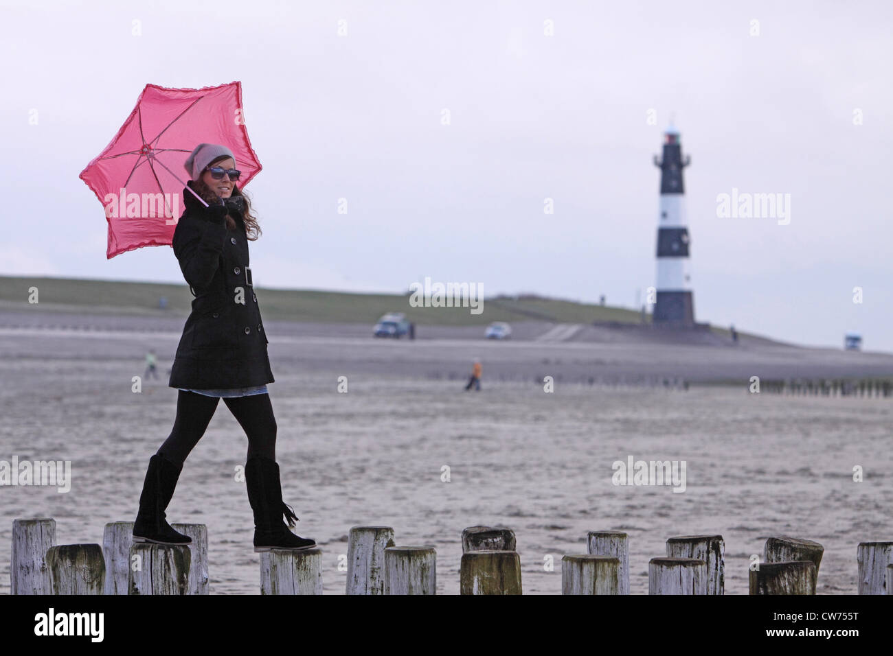 Giovane donna con ombrello rosa in equilibrio su sperone dighe con il faro in background, Paesi Bassi Zeeland, Breskens Foto Stock