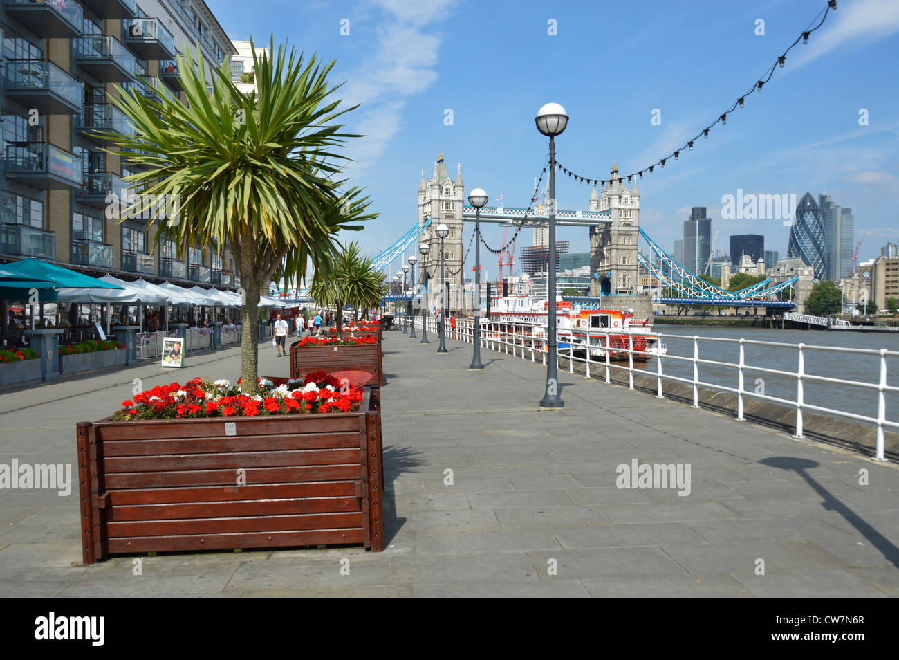 Thames Path Butlers Wharf sul fiume Tamigi viste del Tower Bridge e dello skyline di Londra, ristoranti, tettoie e piantatrici di fiori con alberi di cordyline Londra Regno Unito Foto Stock