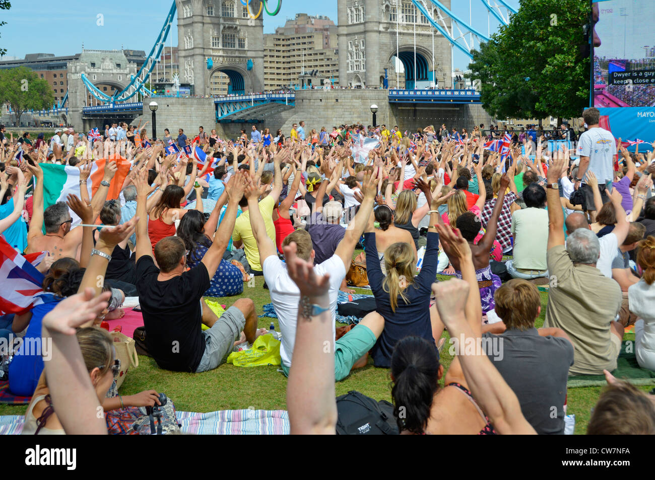 La gente della folla siede sull'erba vista i giochi olimpici di Londra 2012 Il grande schermo della TV mostra le mani in su e il Ponte della Torre ondeggiante Potters Fields Park Southwark Inghilterra Regno Unito Foto Stock