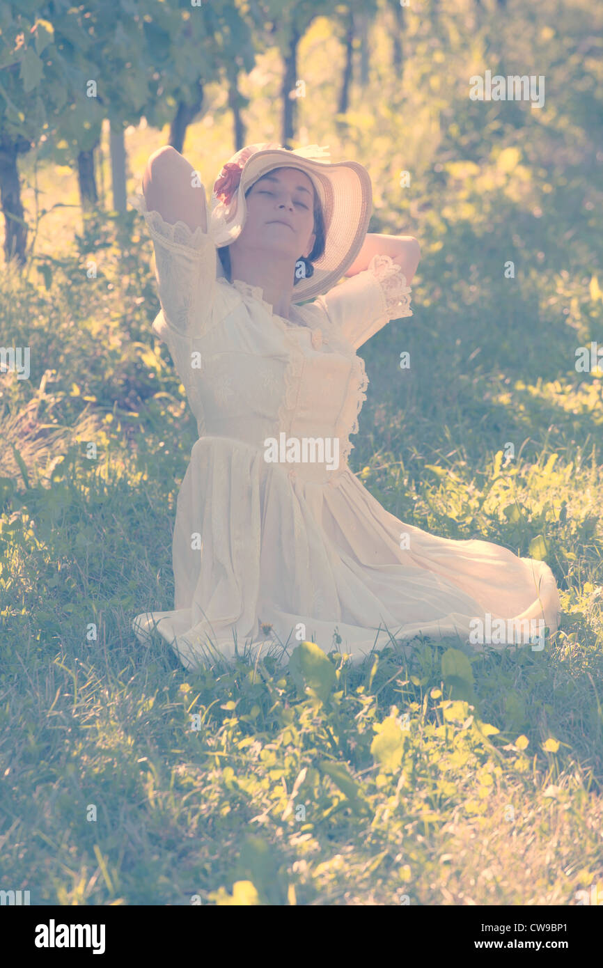 Una donna in bianco abito vittoriano seduto sull'erba tra vigneti e godersi il sole Foto Stock