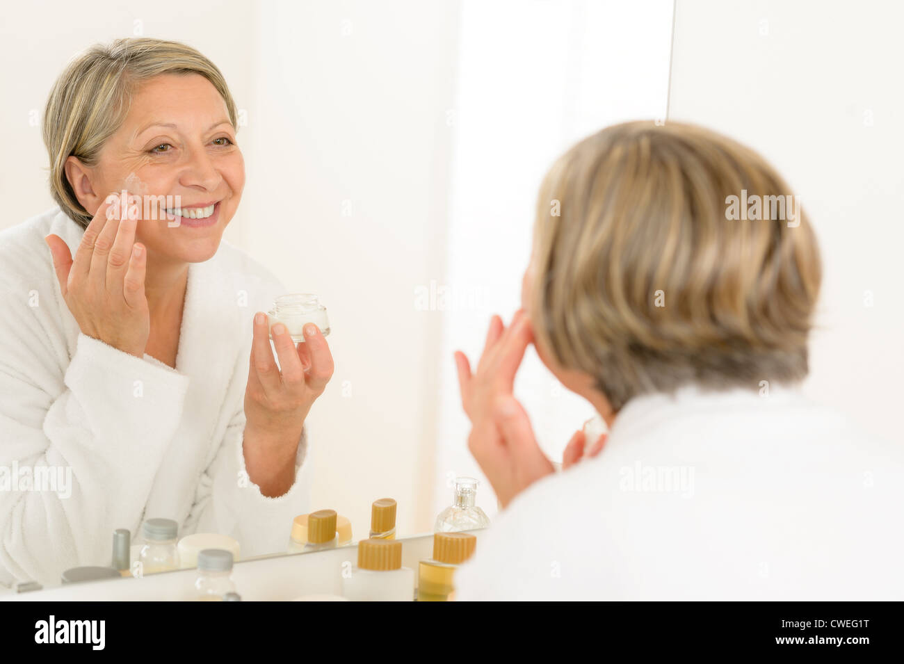 Senior donna che guarda nello specchio del bagno e applicando anti-rughe crema Foto Stock
