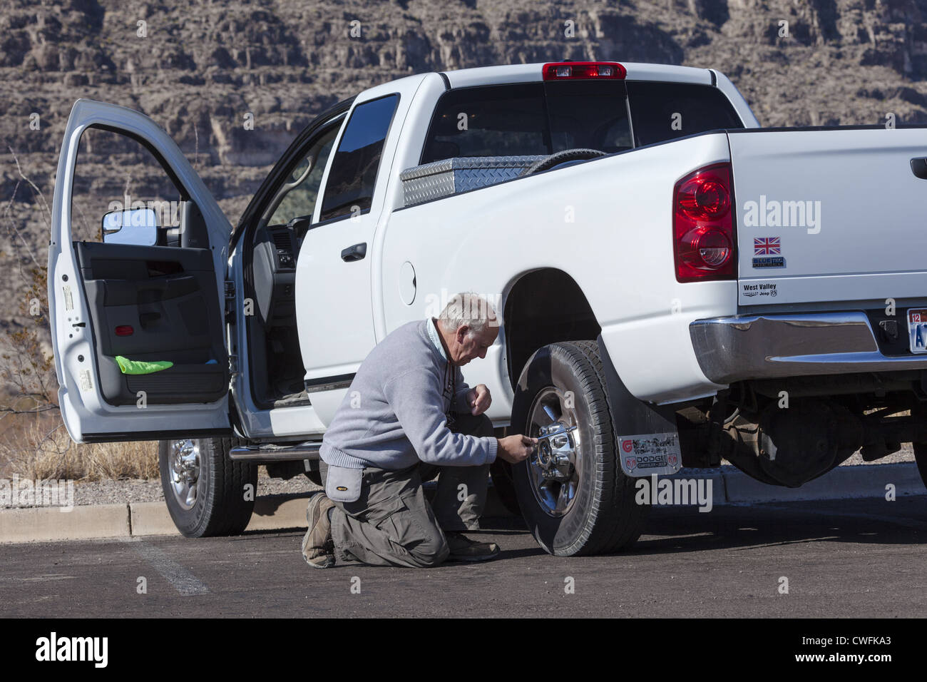 Senior uomo inginocchiato accanto a un bianco pick-up truck controllo della pressione del pneumatico posteriore utilizzando un misuratore di pressione Foto Stock