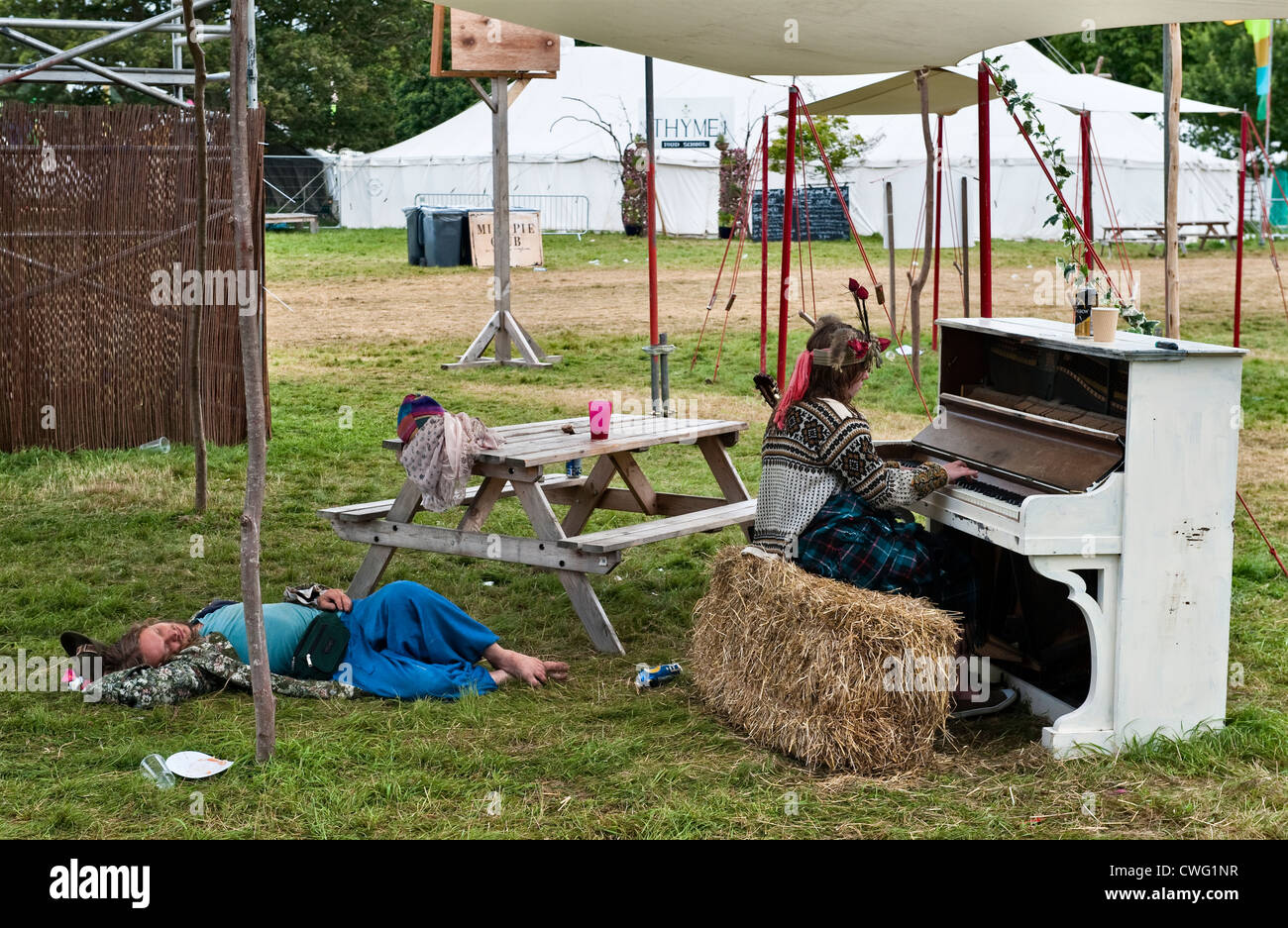 Un goer del festival addormentato a terra la mattina dopo il festival di musica Wilderness, Cornbury, Oxfordshire, UK, mentre un altro suona tranquillamente un pianoforte Foto Stock