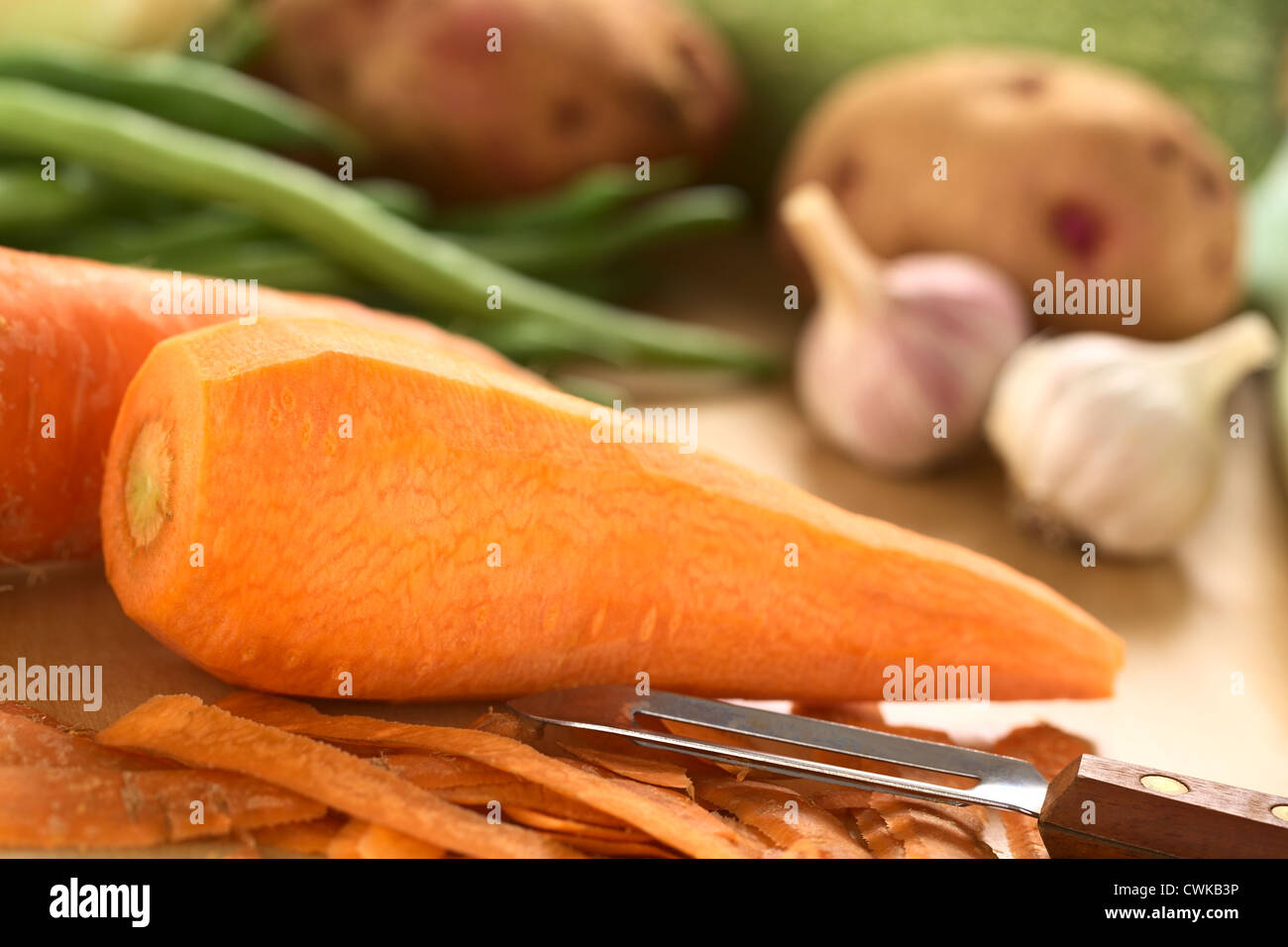 Materie sbucciate la carota con sbucciatore e altri ortaggi (verde fagiolo, patata, aglio) sulla tavola di legno (messa a fuoco selettiva) Foto Stock
