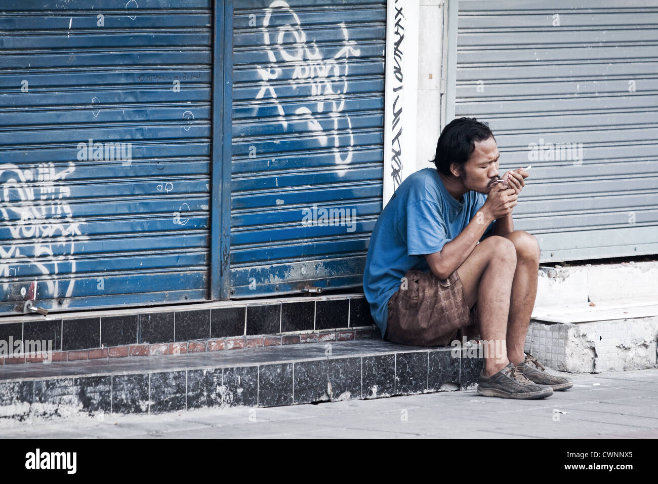 Uomo di illuminazione sat sigarette al di fuori di un negozio a Bangkok. Scena di strada di qualcuno verso il basso e a Bangkok. grintoso sfondo di graffiti sulle serrande del negozio Foto Stock