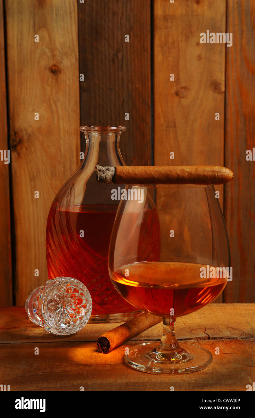 Bicchiere di cognac nella parte anteriore di un decanter di cristallo su di un legno rustico impostazione. Due sigari una accesa e in appoggio sulla parte superiore del vetro. Foto Stock