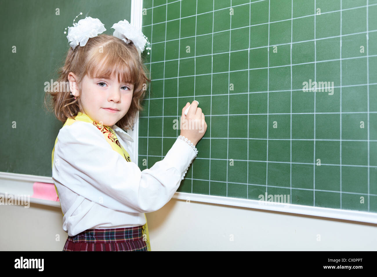 Piccola bellezza ragazza di un allievo di prima classe della scuola elementare in piedi dietro una lavagna verde con Chalk Foto Stock