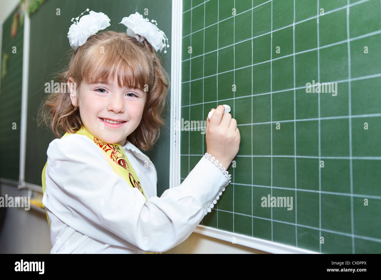 Piccola ragazza felice di un allievo di prima classe della scuola elementare in piedi dietro una lavagna verde con Chalk Foto Stock