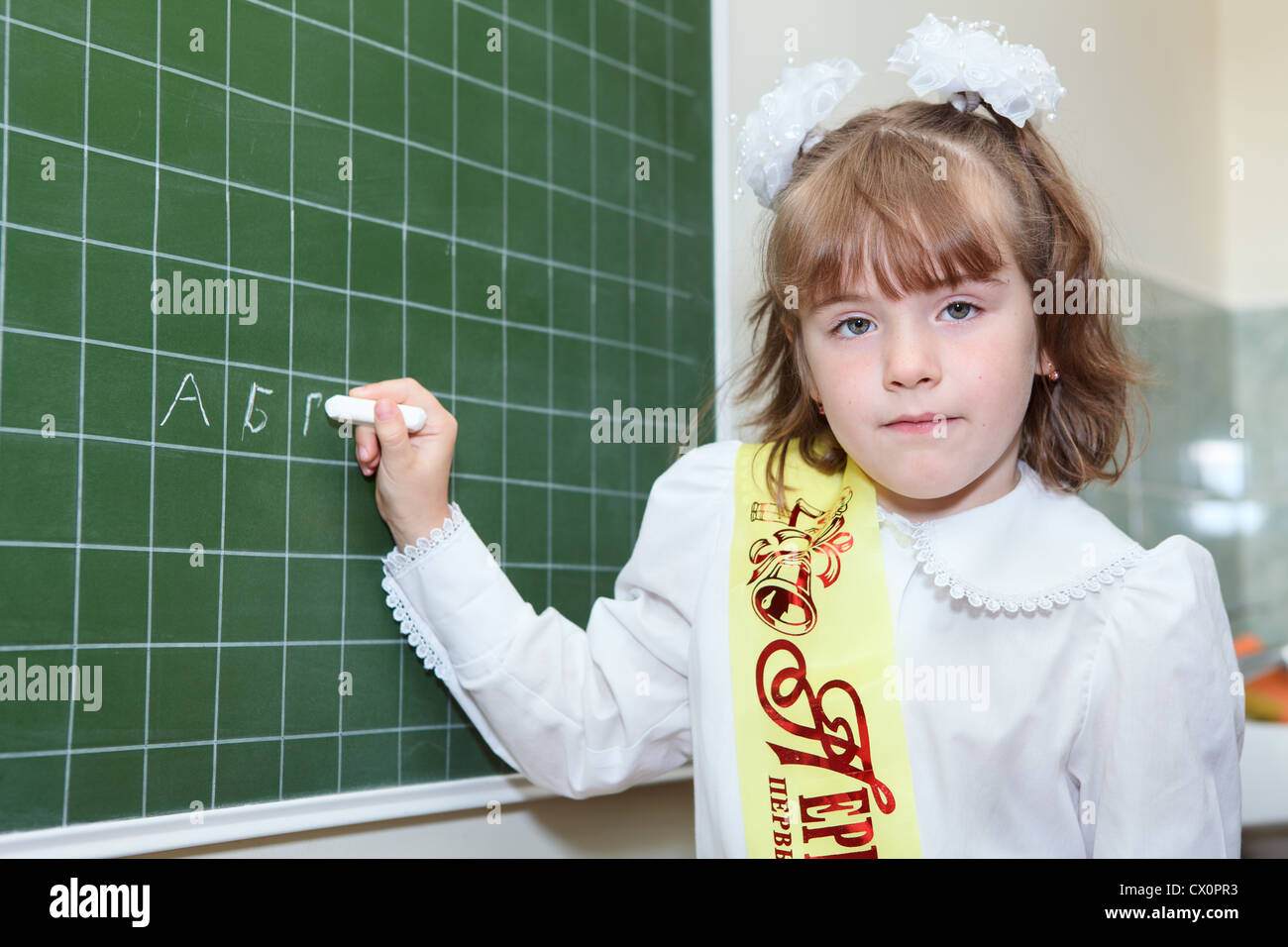 Bambina di un allievo di prima classe della scuola elementare in piedi dietro una lavagna verde con il gesso e la scrittura di una lettera Foto Stock