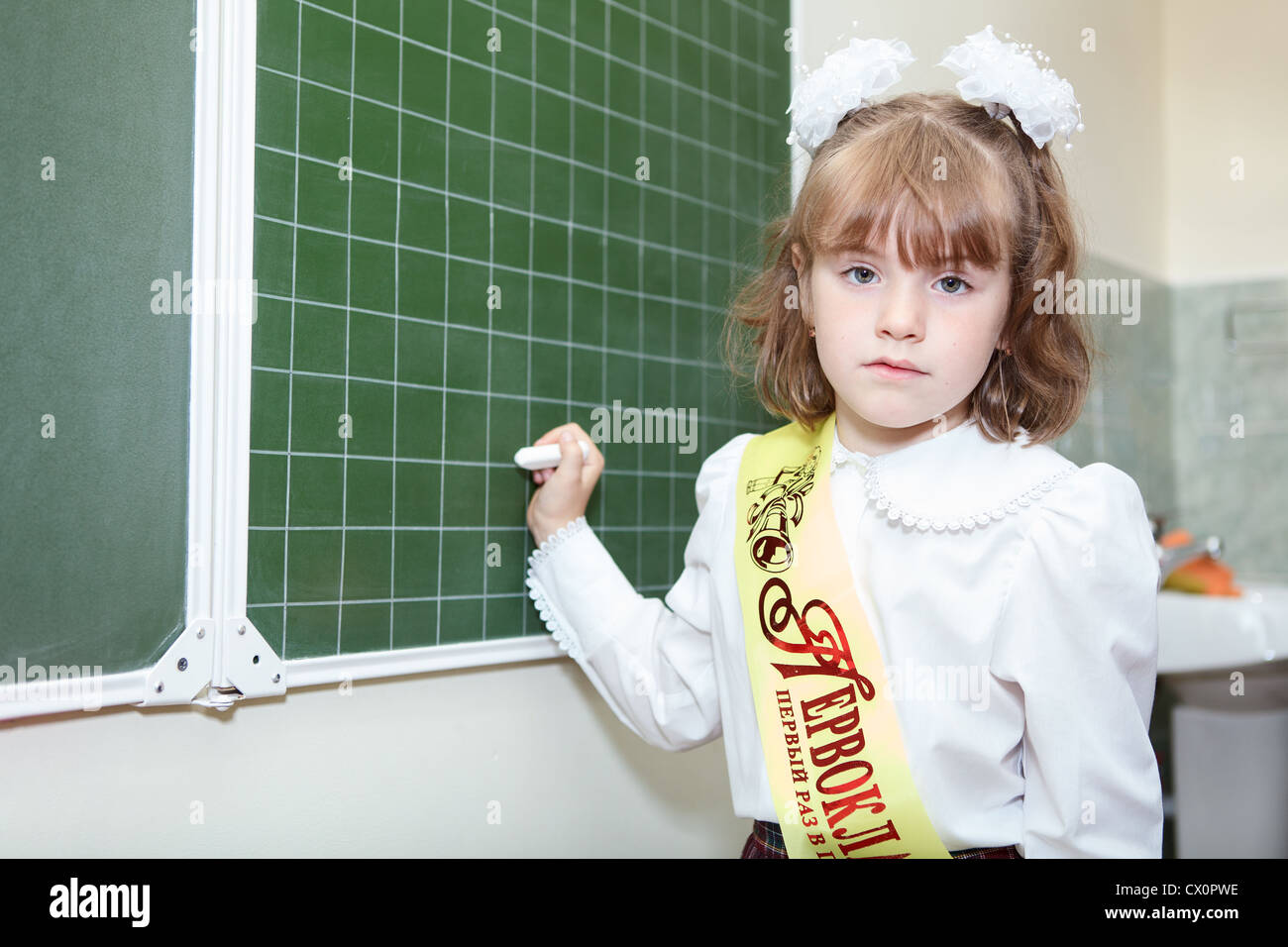 Poco ragazza russa di un allievo di prima classe della scuola elementare in piedi dietro una lavagna verde con Chalk Foto Stock