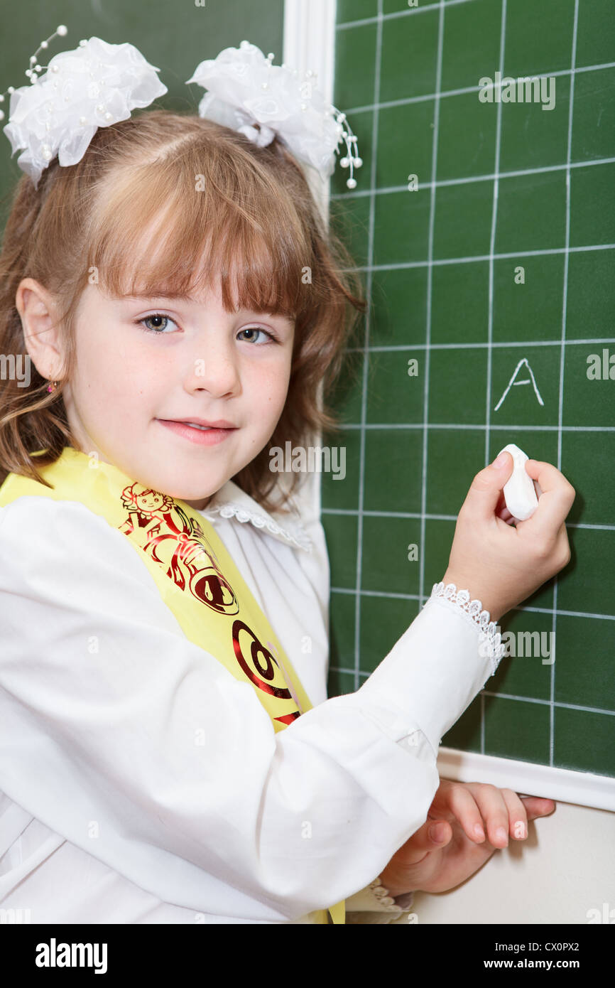 Piccola ragazza di un allievo di prima classe della scuola elementare in piedi dietro una lavagna verde con il gesso e la scrittura di una lettera Foto Stock