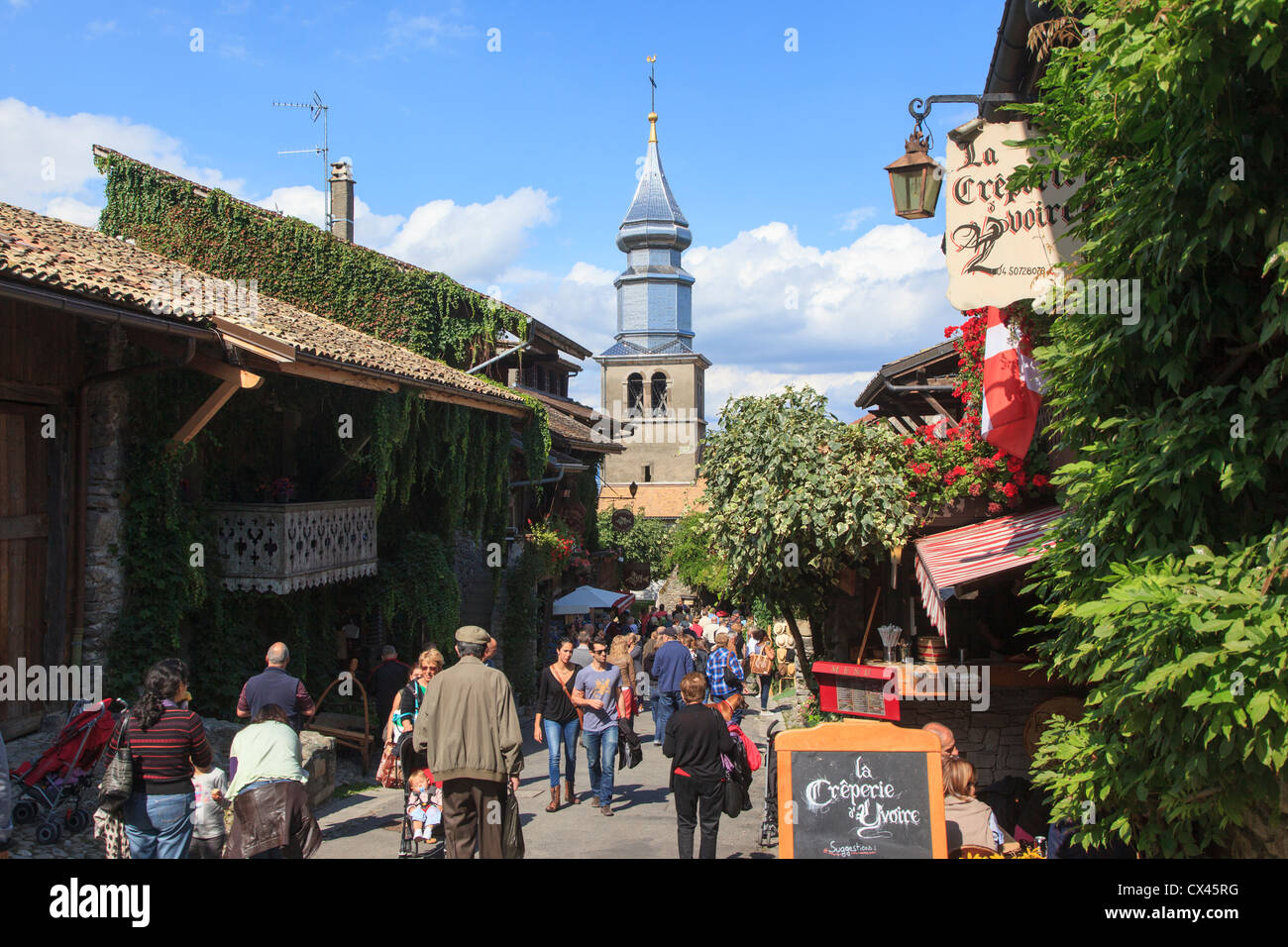 Tourist riempito street a Yvoire Francia con la lucida il campanile della chiesa di St Pancras in background Foto Stock
