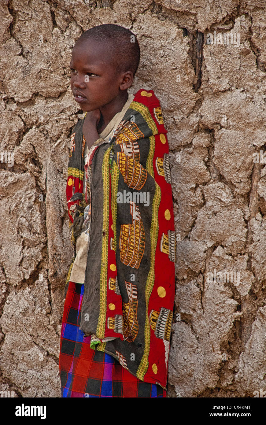Masai Bambino, indossando abiti tradizionali, in piedi contro un muro di fango in un villaggio Masai Mara, Kenya, Africa Foto Stock