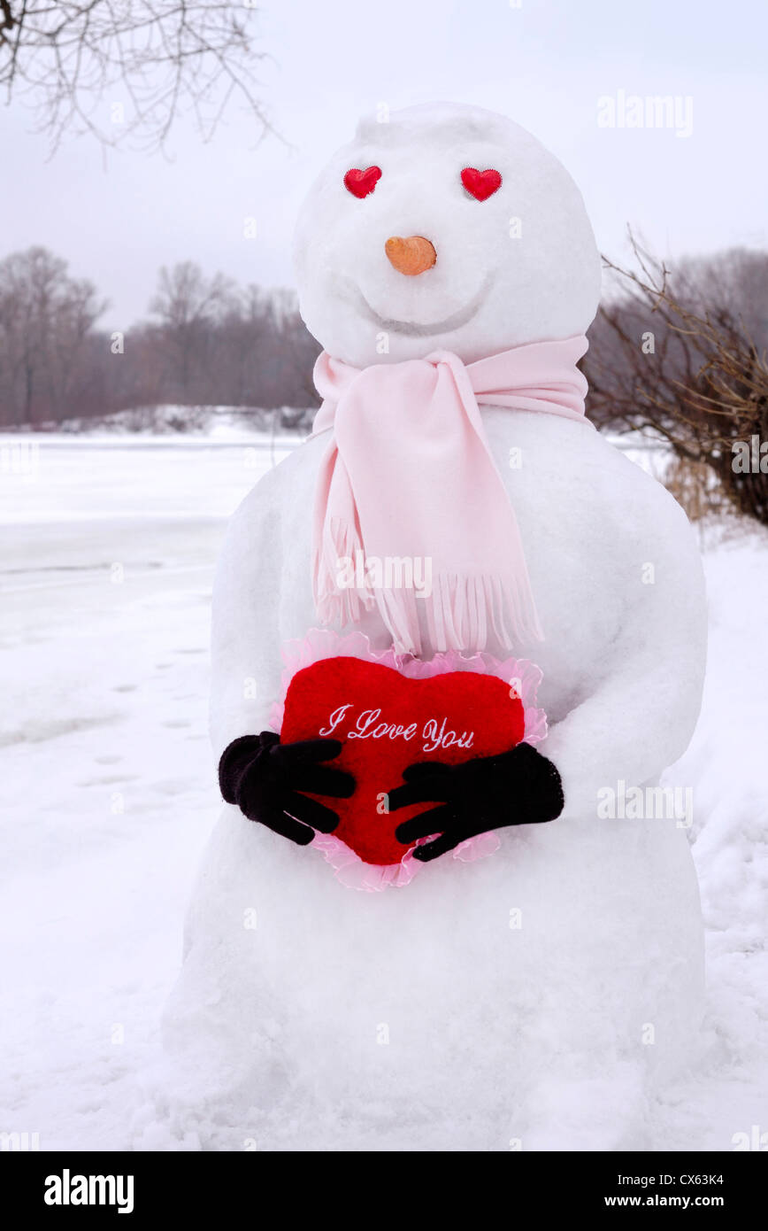Sorridente pupazzo di neve con la a forma di cuore gli occhi tenendo un cuore rosso con Ti Amo scritto su di esso nelle sue mani. Il giorno di San Valentino concetto Foto Stock