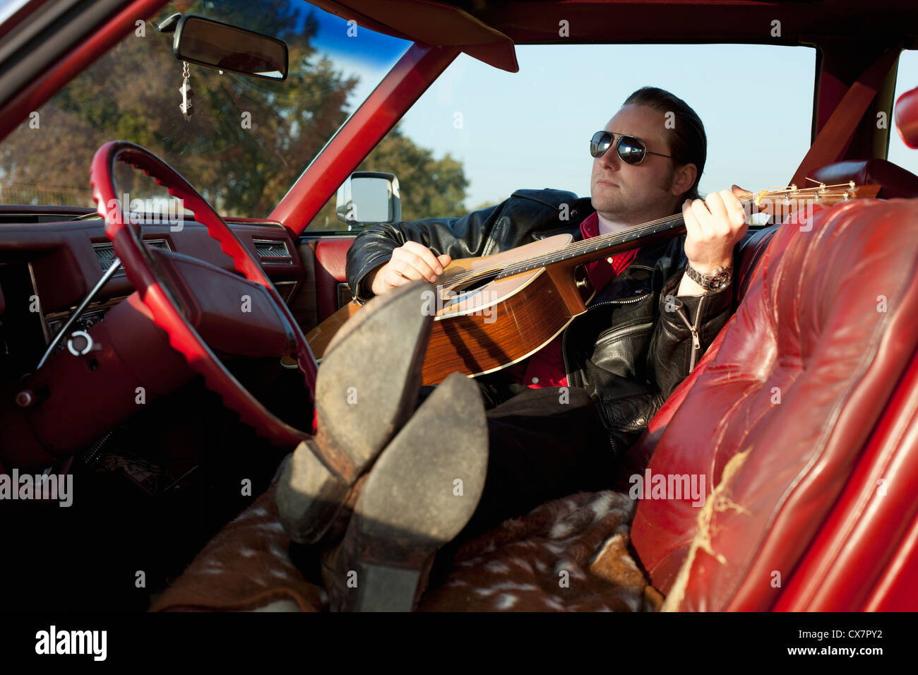 Un rockabilly guy suonando una chitarra acustica mentre è seduto nella sua auto d'epoca Foto Stock
