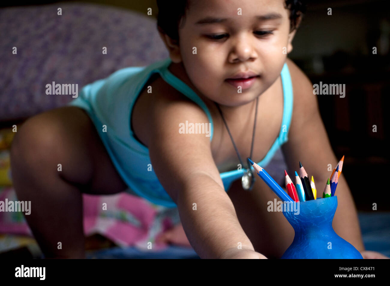 Una ragazza indiana sta giocando con le matite colorate tenute in un piccolo vaso blu seduta sul suo letto. Foto Stock