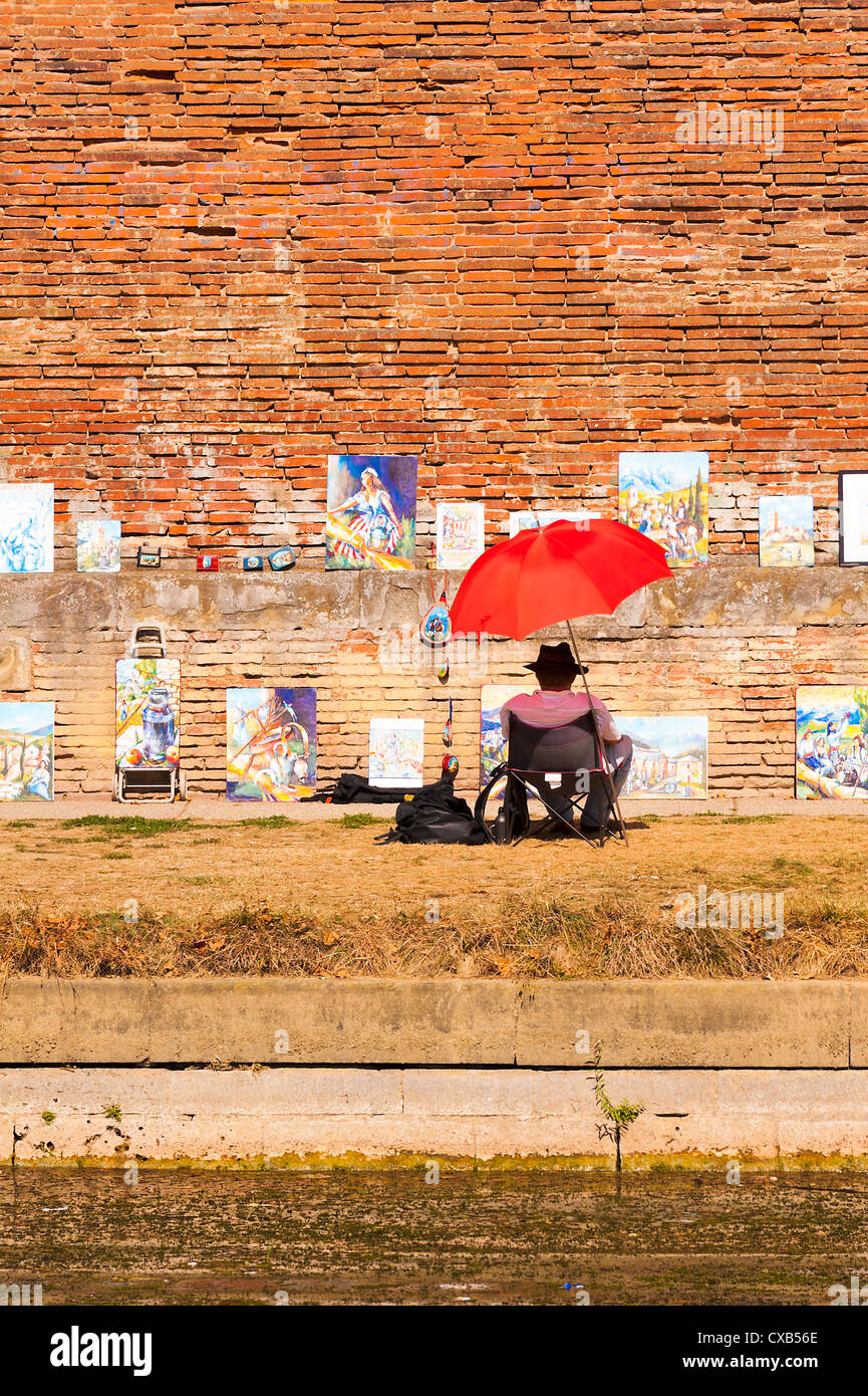 Artista seduto sotto un ombrello rosso in ombra vendere dipinti sulla banca del fiume Garonne Toulouse Midi-Pirenei Francia Foto Stock