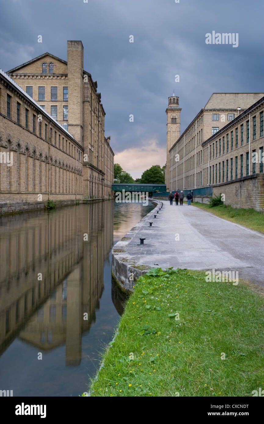 4 persone a piedi sulla strada alzaia del Leeds Liverpool canal dagli storici, impressionante Salts Mill (edifici riflessa nell'acqua) - Saltaire, nello Yorkshire, Inghilterra, Regno Unito Foto Stock