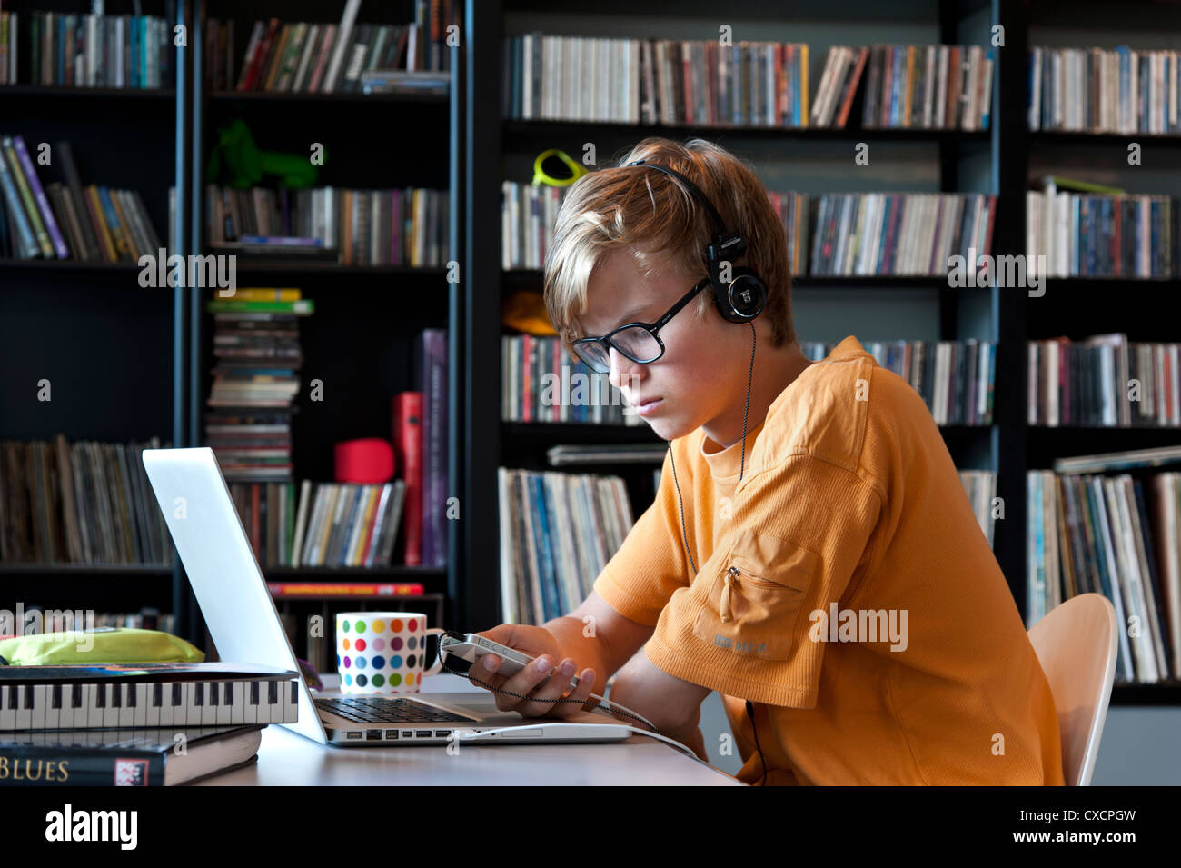 Studiosa rilassato scuola teenage boy 14-16 anni di indossare le cuffie utilizzando il proprio Apple iPhone smartphone e computer portatili in musica libreria di studio Foto Stock