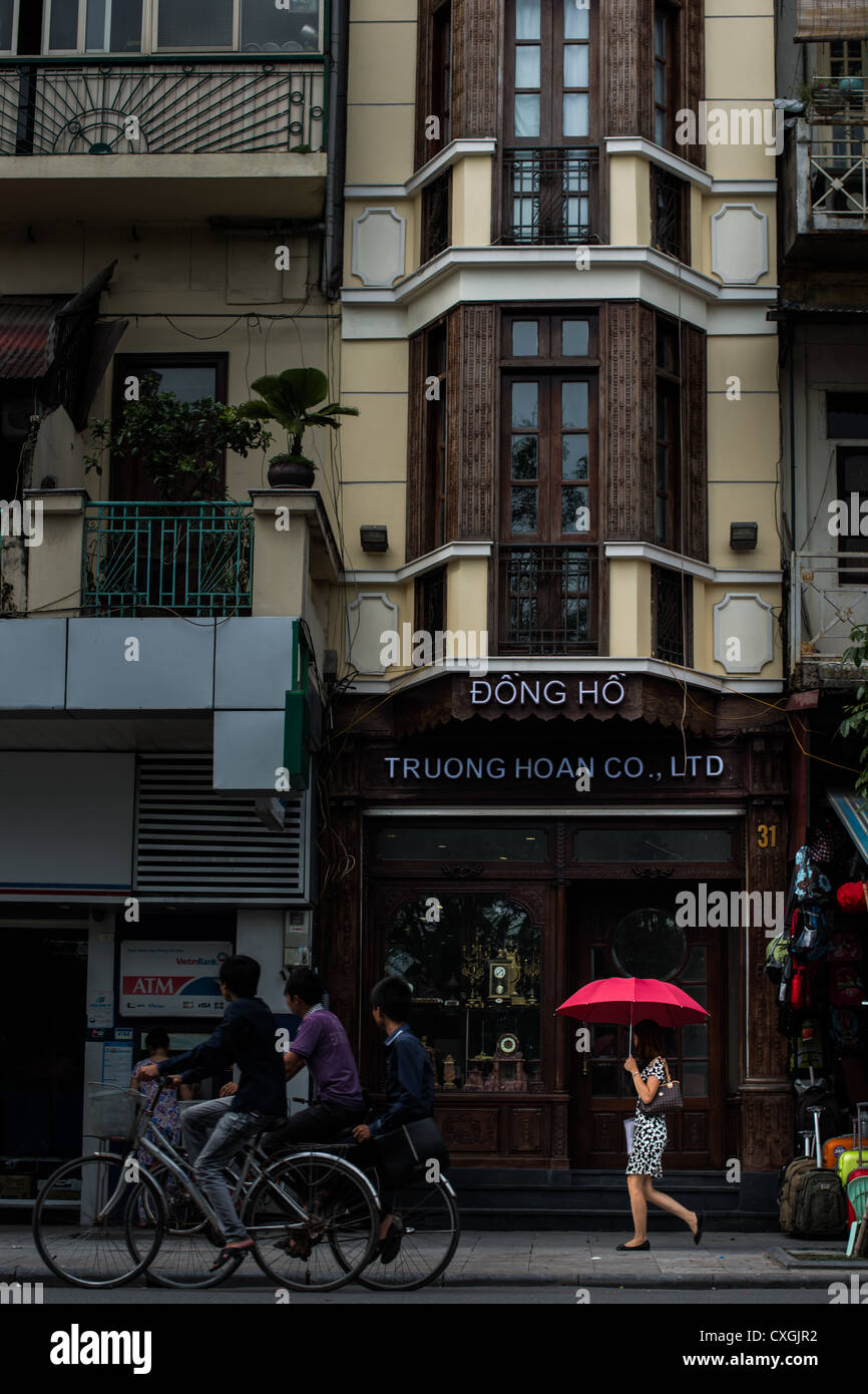 Donna con ombrello rosso bagnato in un fugace raggio di sole passeggiate da un negozio tradizionale nella parte anteriore della vecchia di Hanoi Foto Stock