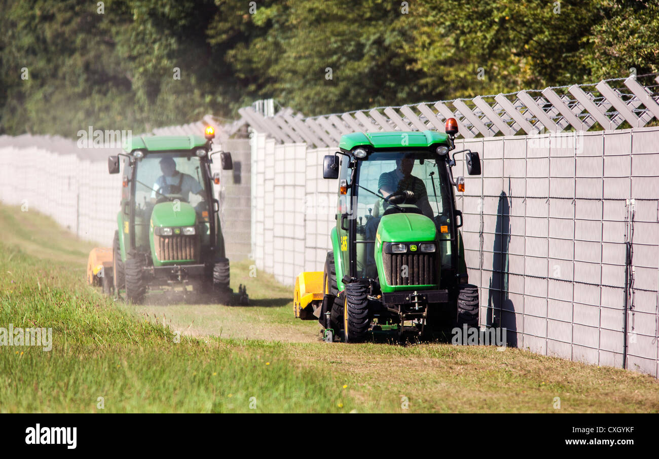 Trattori Falciare il prato lungo una recinzione di sicurezza, toped con filo spinato per proteggere un'area. Foto Stock