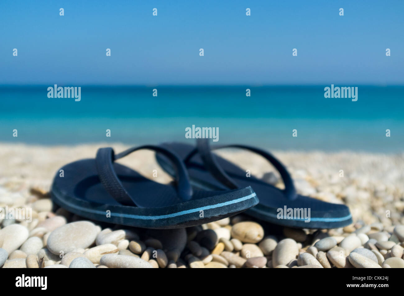 Una coppia di plastica blu flip flop su una spiaggia di ciottoli con un mare blu in background in soft focus. Foto Stock