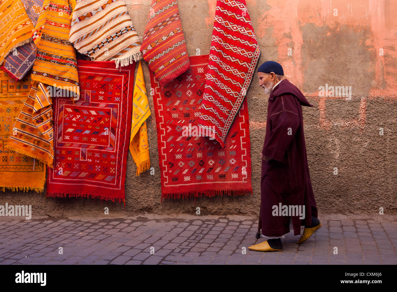 Uomo marocchino passato a piedi di tappeti sospesi alla parete indossando djellaba e babouch pantofole, Marrakech, Marocco Foto Stock