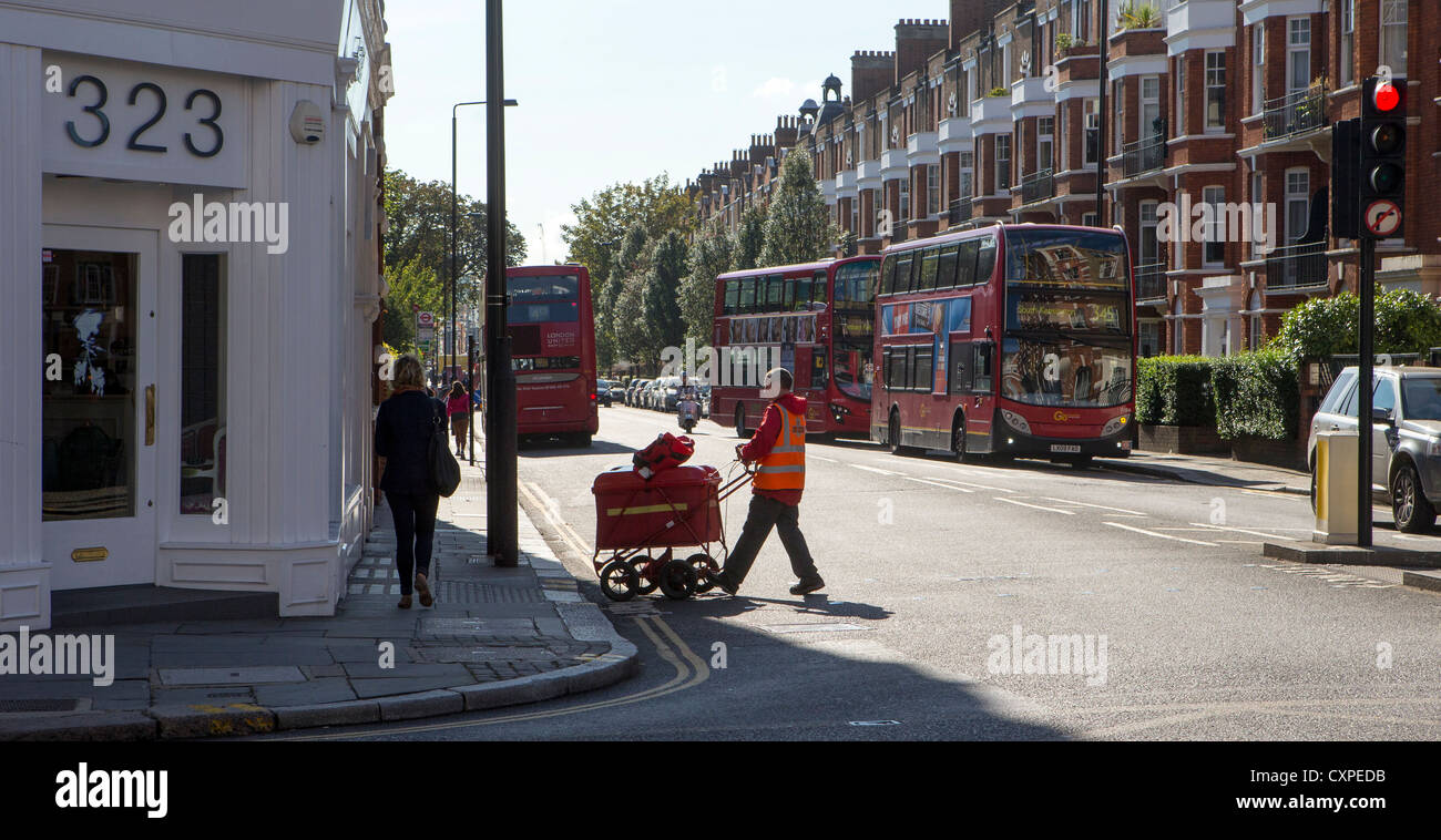 Un postino Londra attraversando la strada e wheeling il suo email carrello / carrello di fronte a lui, con autobus rossi di Londra dietro. Foto Stock