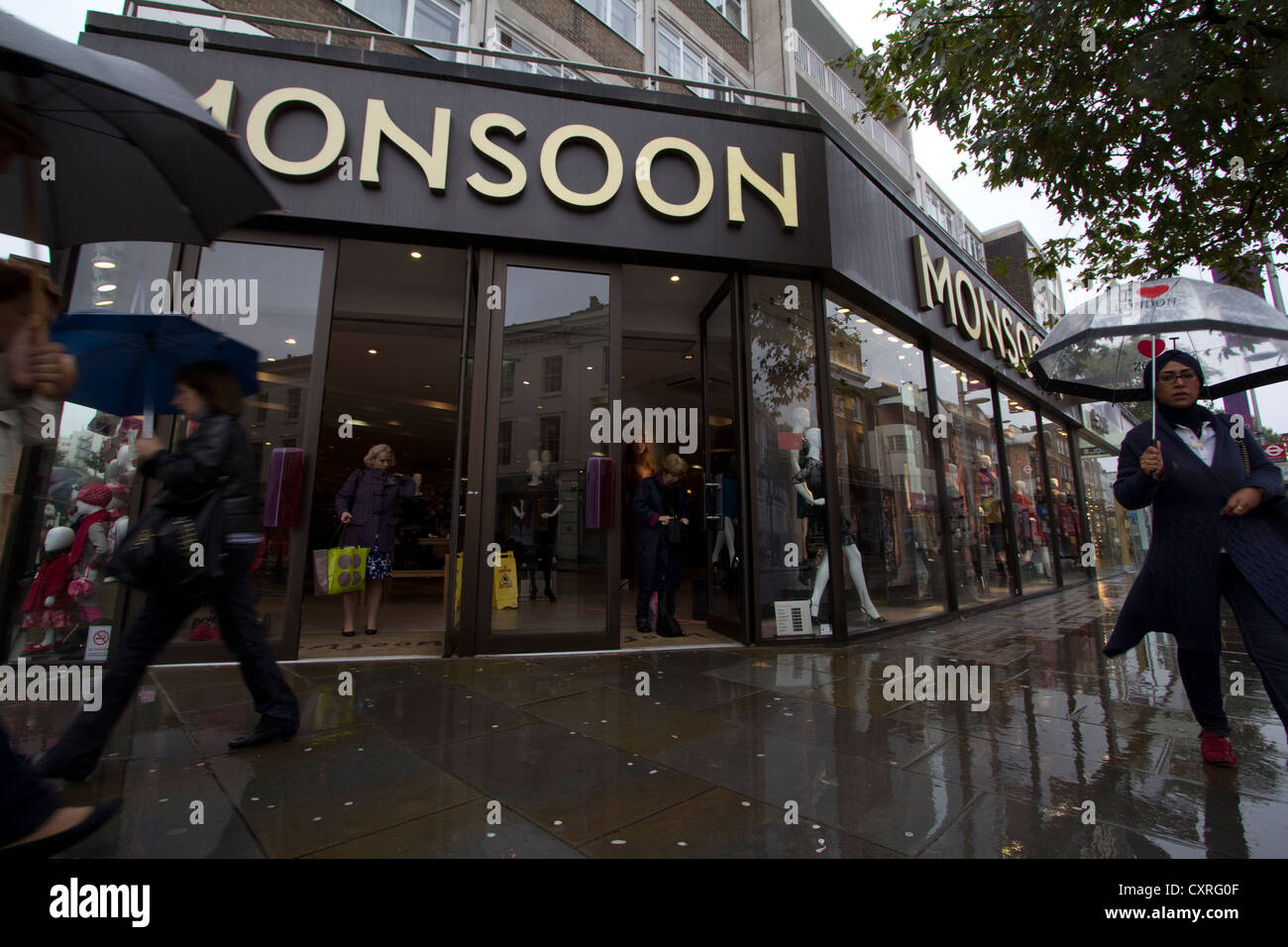 Durante un temporale, le persone con ombrelloni passano davanti al negozio di moda Monsoon a Londra, Regno Unito, catturando l'essenza dello shopping nelle strade principali britanniche, il clima stagionale e il fascino duraturo dei marchi di moda femminili contemporanei Foto Stock