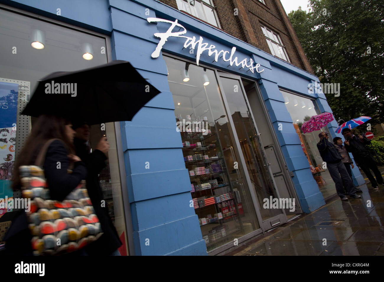 Gli amanti dello shopping con ombrelloni passano davanti al punto vendita Paperchase su King's Road, Londra, durante una giornata di pioggia, illustrando la cultura dello shopping nelle strade principali britanniche, il clima inclemente e la resilienza del traffico pedonale in una delle destinazioni più popolari di Londra Foto Stock