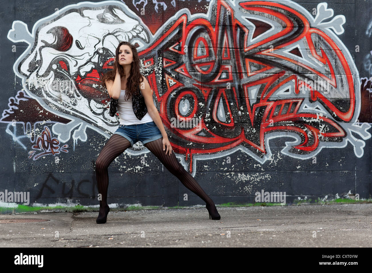 Giovane donna in un top bianco, denim hot pants, calze nere e tacchi alti che pongono di fronte a un muro di graffiti Foto Stock