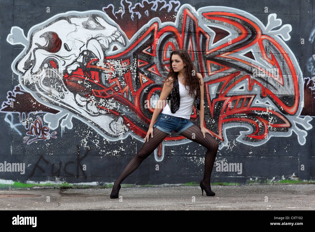Giovane donna in un top bianco, denim hot pants, calze nere e tacchi alti che pongono di fronte a un muro di graffiti Foto Stock