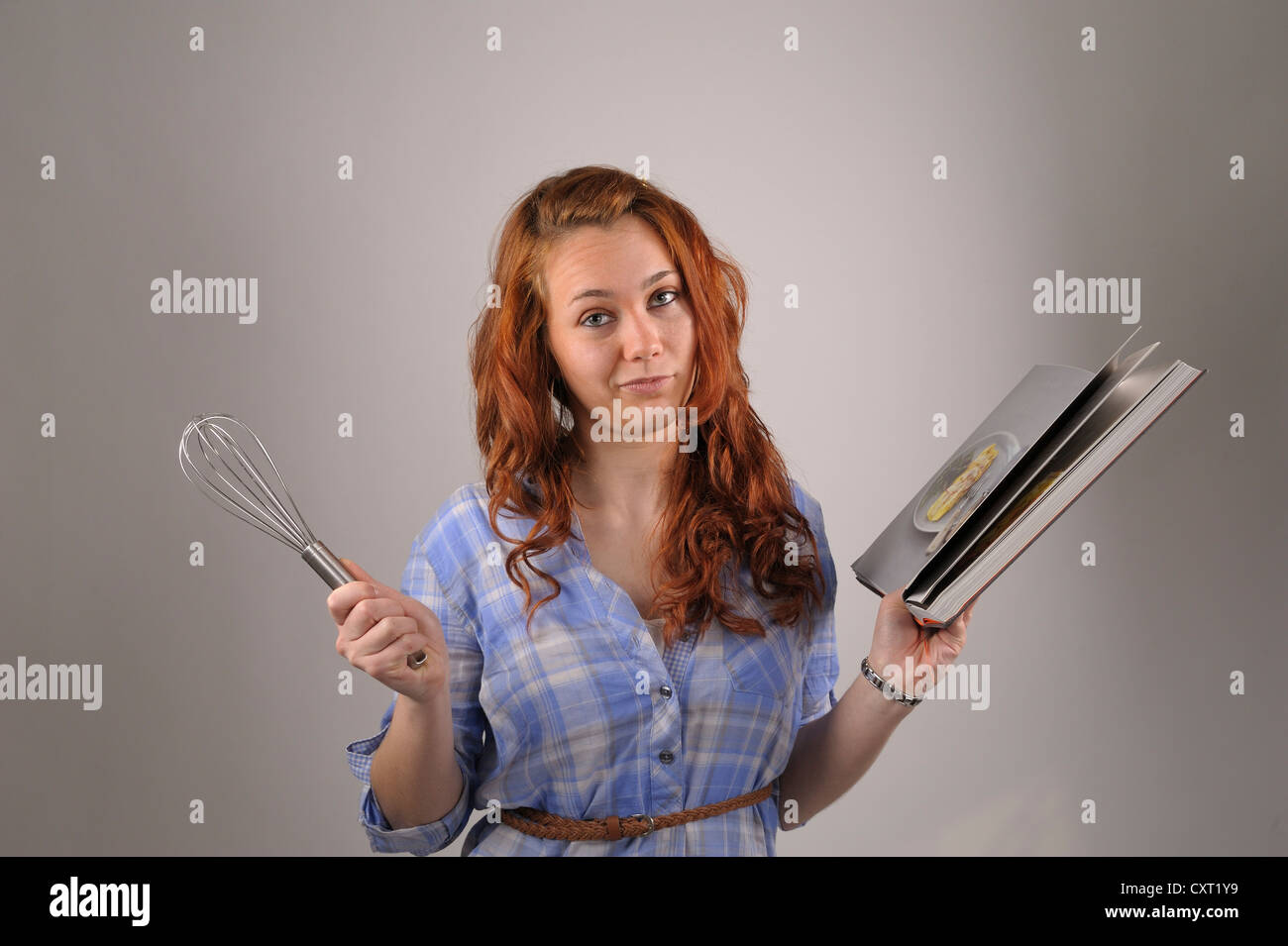 Giovane donna con i capelli rossi tenendo una frusta e un libro di cucina nelle sue mani Foto Stock