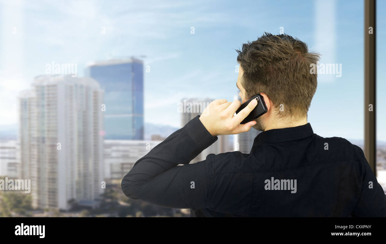 Giovane imprenditore parlando sul suo telefono cellulare di fronte a una finestra una skyline sul retro, visto da dietro Foto Stock
