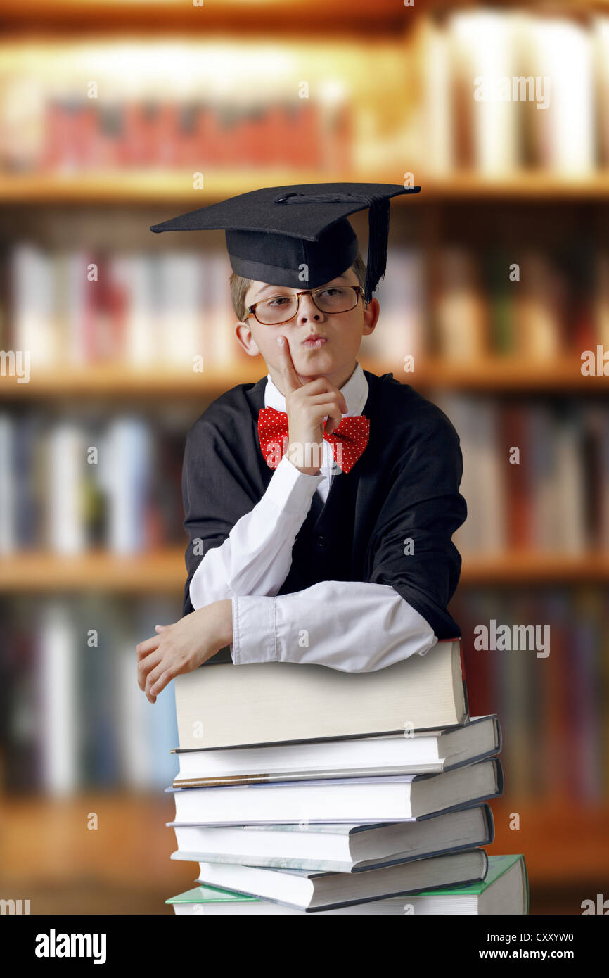 Riflessivo cercando schoolboy indossando un cappello di graduazione e appoggiato su di un pile di libri in una libreria Foto Stock