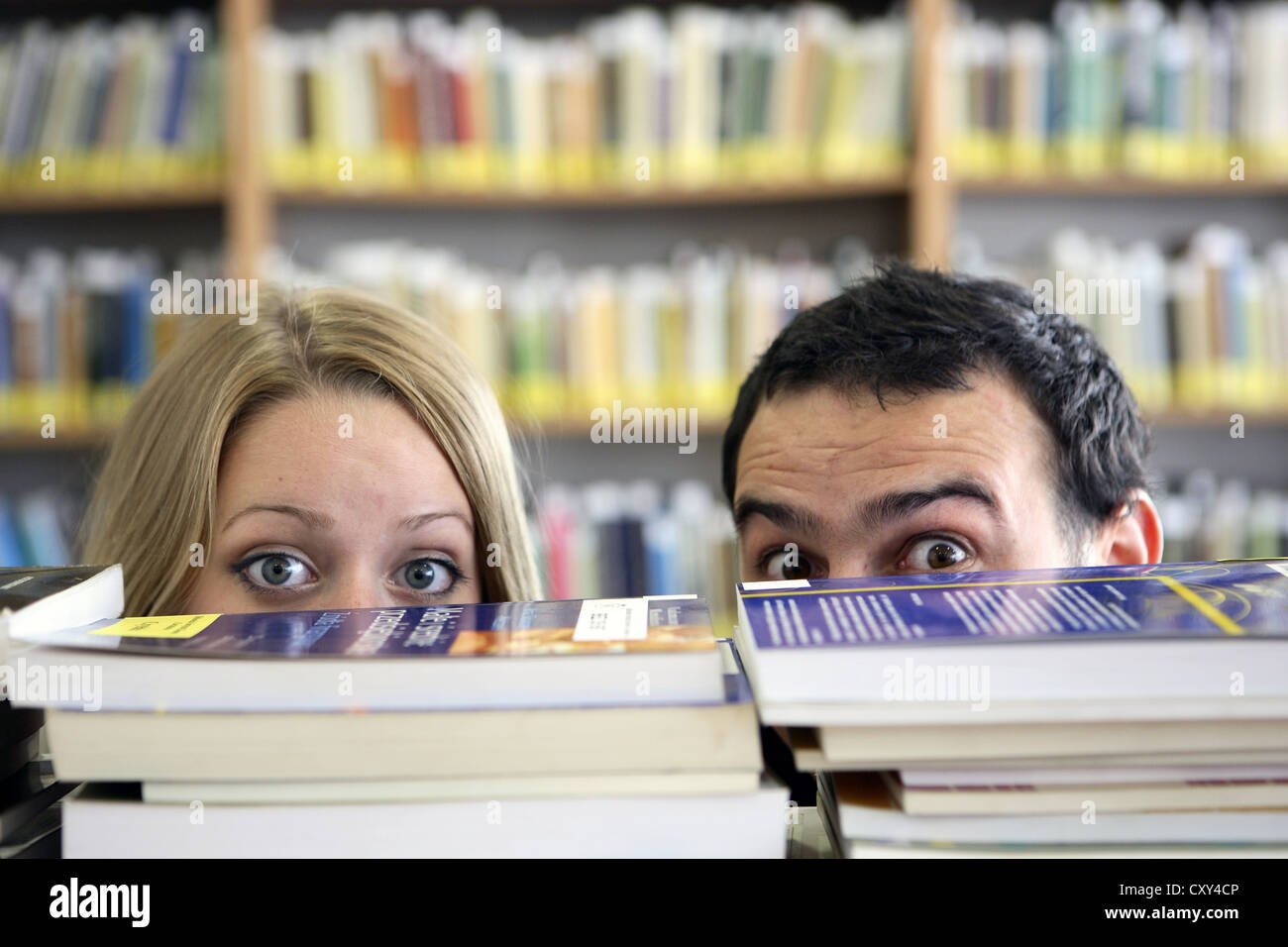 Gli studenti, coppia, guardando fuori da dietro pile di libri, biblioteca universitaria Foto Stock