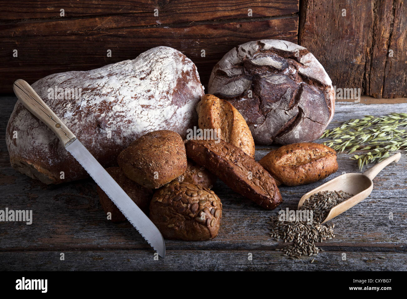 Forme di pane, panini e un coltello per pane di segale, grano e spighe di grano su un rustico di una superficie di legno Foto Stock