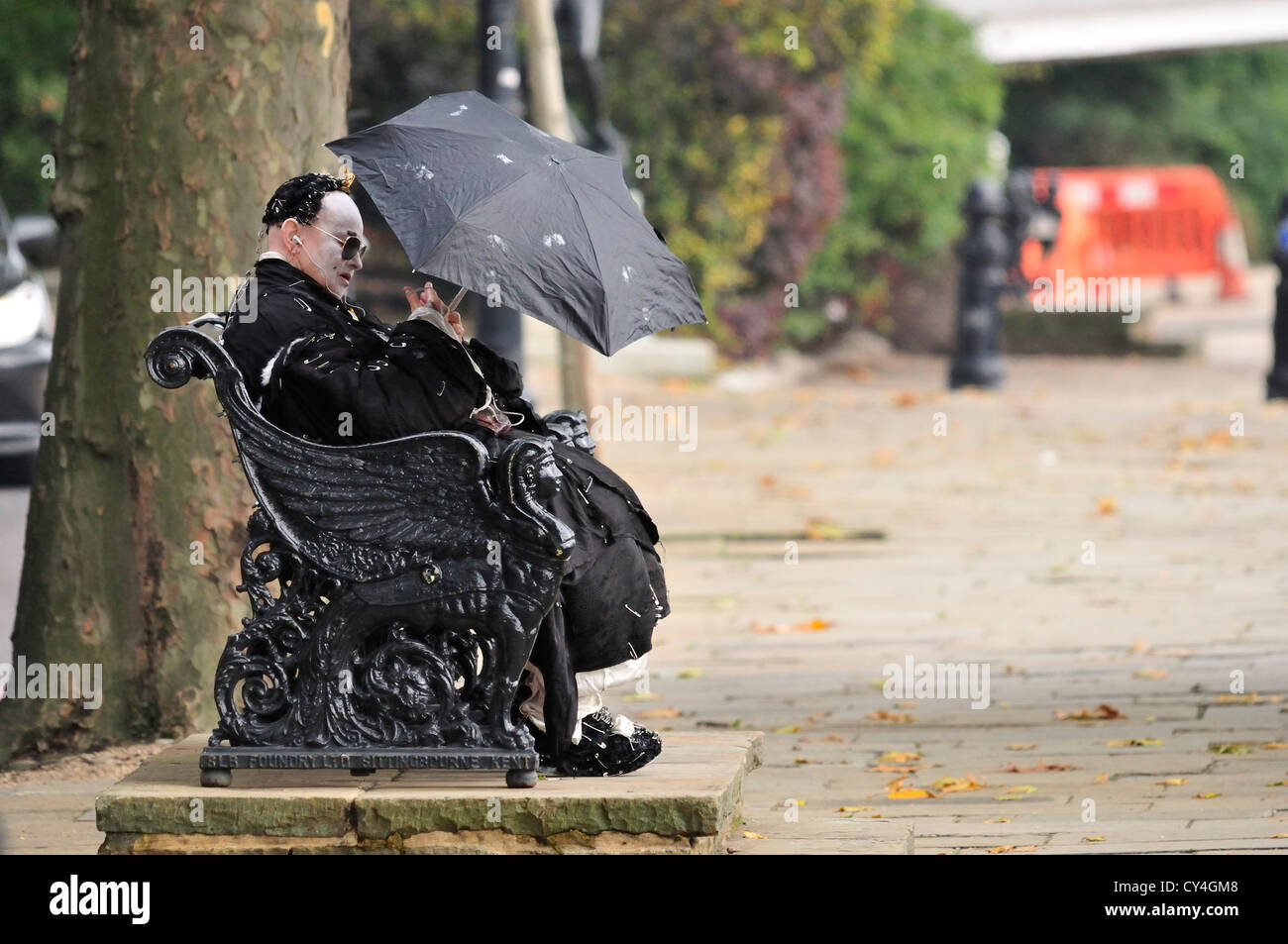 Londra, Inghilterra, Regno Unito. L'uomo vestito di nero con faccia bianca e coperti di spine di sicurezza, con un ombrello in una giornata di sole Foto Stock