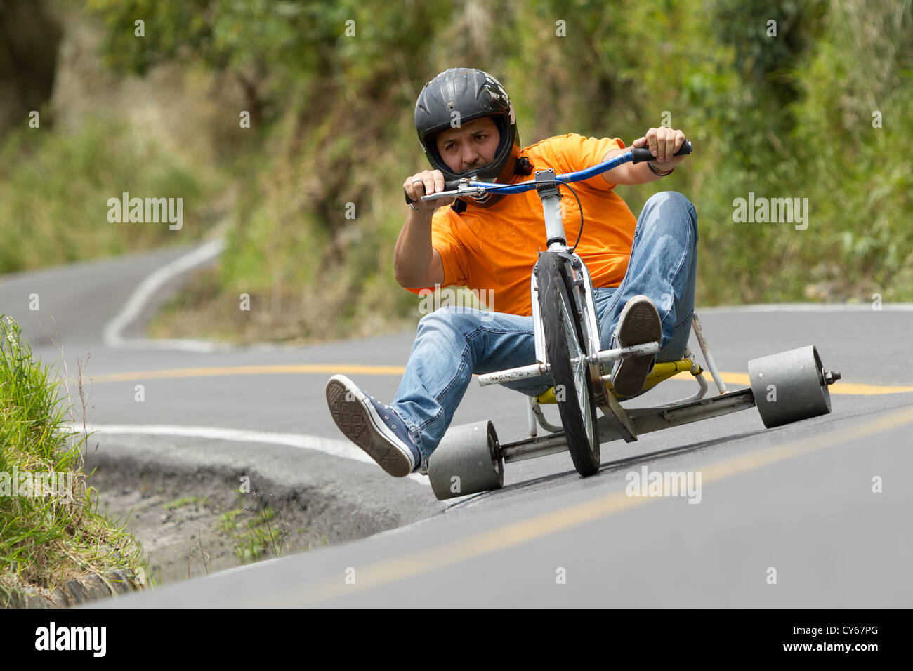 Un uomo che indossa un casco nero, una t-shirt arancione e un jeans blu guida uno scooter personalizzato a tre ruote lungo una strada asfaltata e tortuosa. È seduto con i piedi estesi, afferrando il manubrio, e. Foto Stock