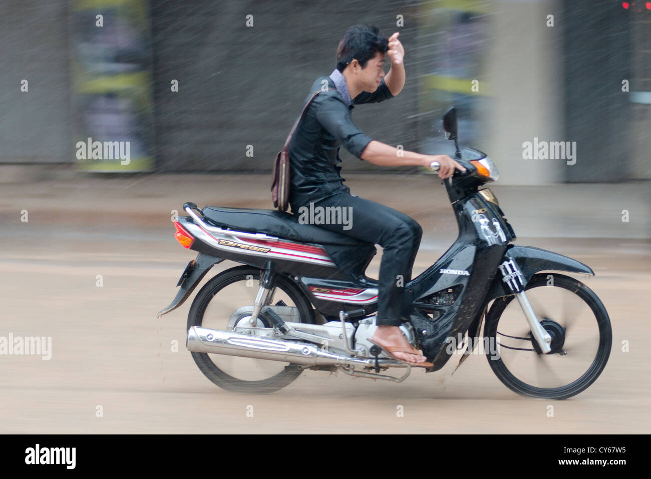 Un giovane uomo di battaglie attraverso la guida sotto la pioggia sulla sua moto nel Sisophon, Cambogia Foto Stock