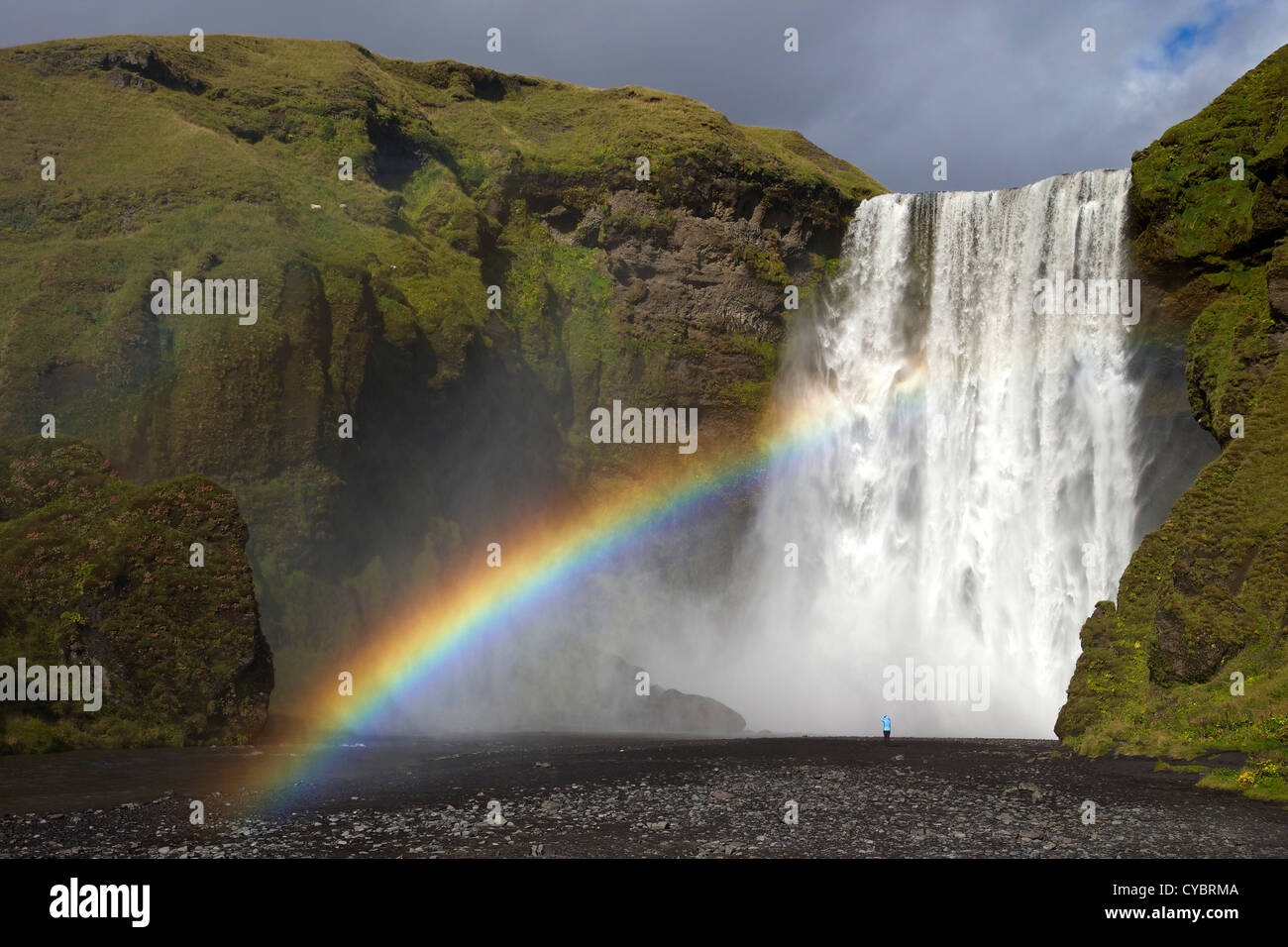 Una persona in piedi vicino alla cascata Skogafoss con arcobaleno nel sole estivo, South Coast, Islanda Foto Stock