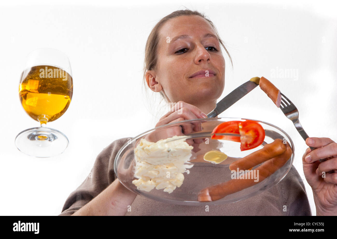 Giovane donna è mangiare a un tavolo di vetro. Salsicce viennesi con insalata di patate, senape e un bicchiere di birra. Foto Stock