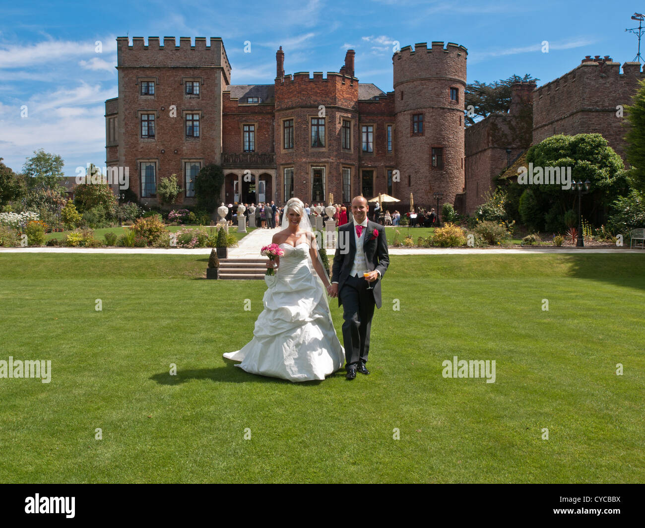 Un Recentemente sposato sposa e lo sposo passeggiata mano nella mano attraverso le impressionanti giardini di un vecchio castello inglese sul loro giorno di nozze Foto Stock