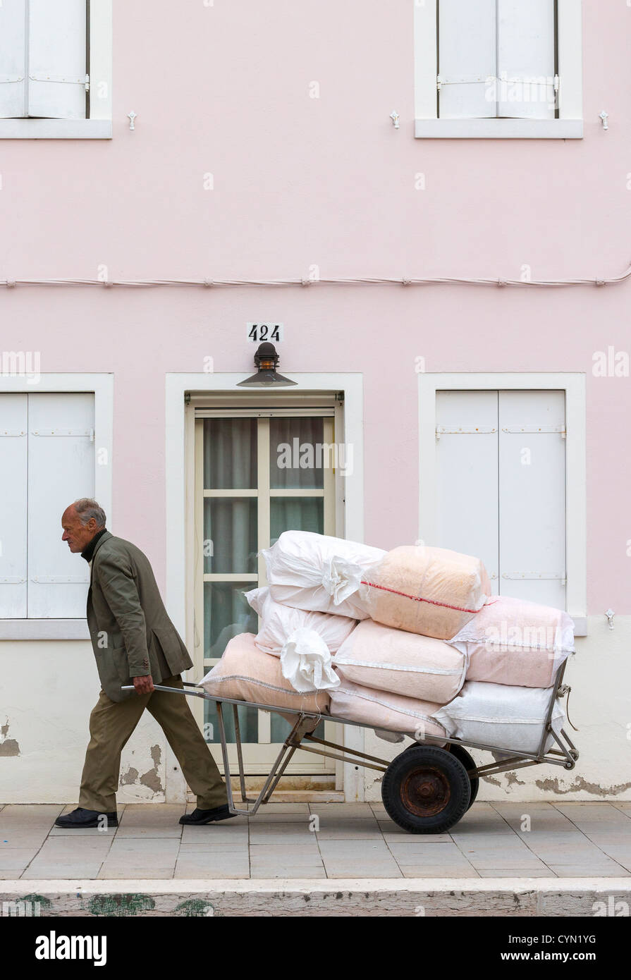 Un vecchio uomo tira un carrello carico di bianco e rosa sacchi a piedi nella parte anteriore di una casa rosa Foto Stock
