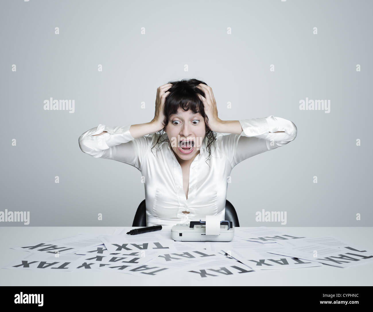 Giovane donna sorpresa alla sua scrivania spaventati da tasse grida tenendo le mani sul suo capo Foto Stock