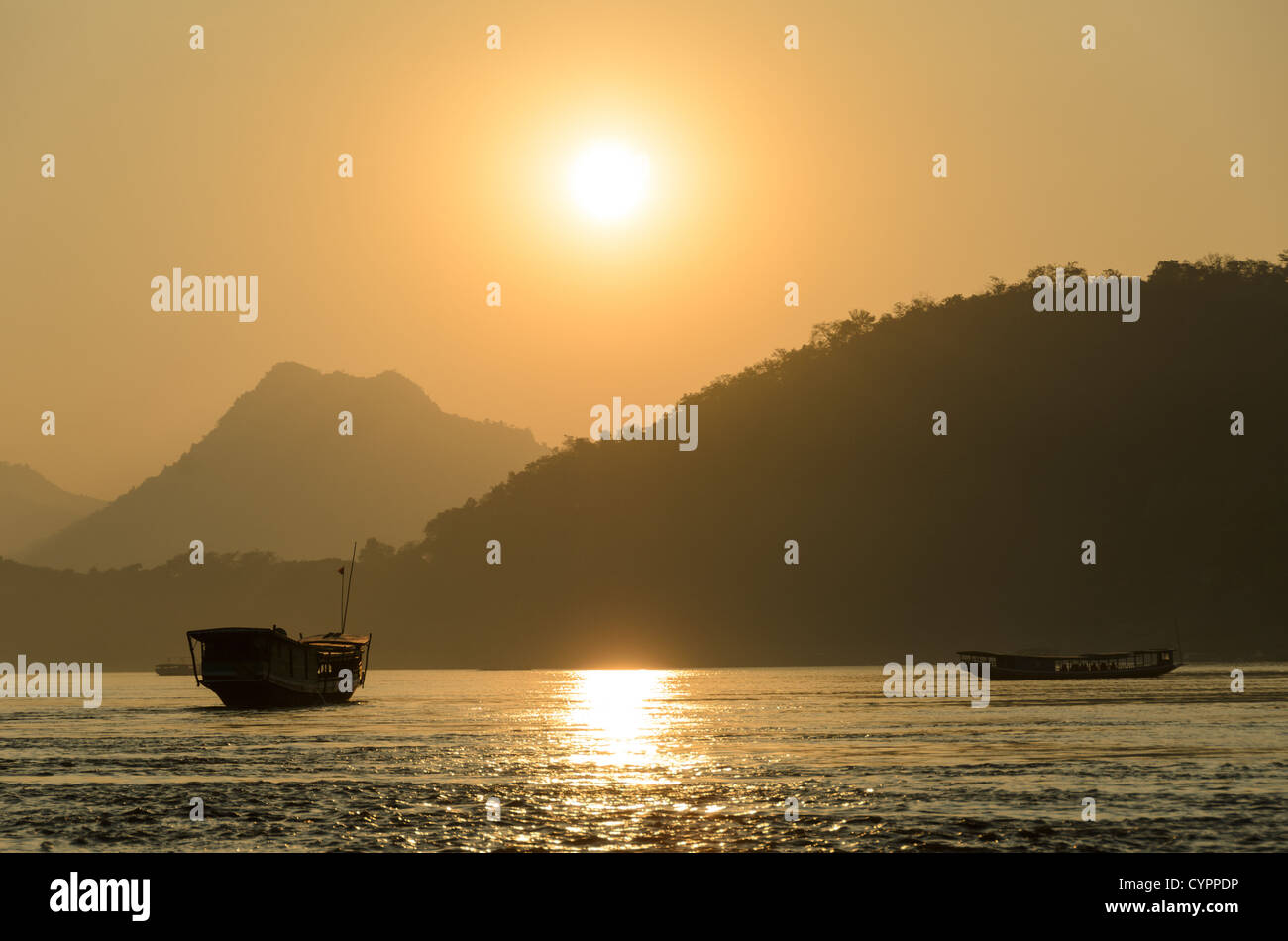 Il fiume Mekong batte al tramonto Luang Prabang Laos // LUANG PRABANG, Laos - il fiume Mekong scorre oltre questa città patrimonio dell'umanità dell'UNESCO mentre le tradizionali barche in legno attraversano il corso d'acqua in lontananza. La luce dorata filtra attraverso la foschia atmosferica durante il tramonto, creando sagome delle montagne circostanti. Luang Prabang si trova alla confluenza dei fiumi Mekong e Nam Khan nel Laos settentrionale. La città è stata la capitale reale del Regno del Laos fino al 1975 e rimane un importante centro culturale. Il trasporto fluviale tradizionale continua a svolgere un ruolo fondamentale nel commercio locale e nella vita quotidiana Foto Stock